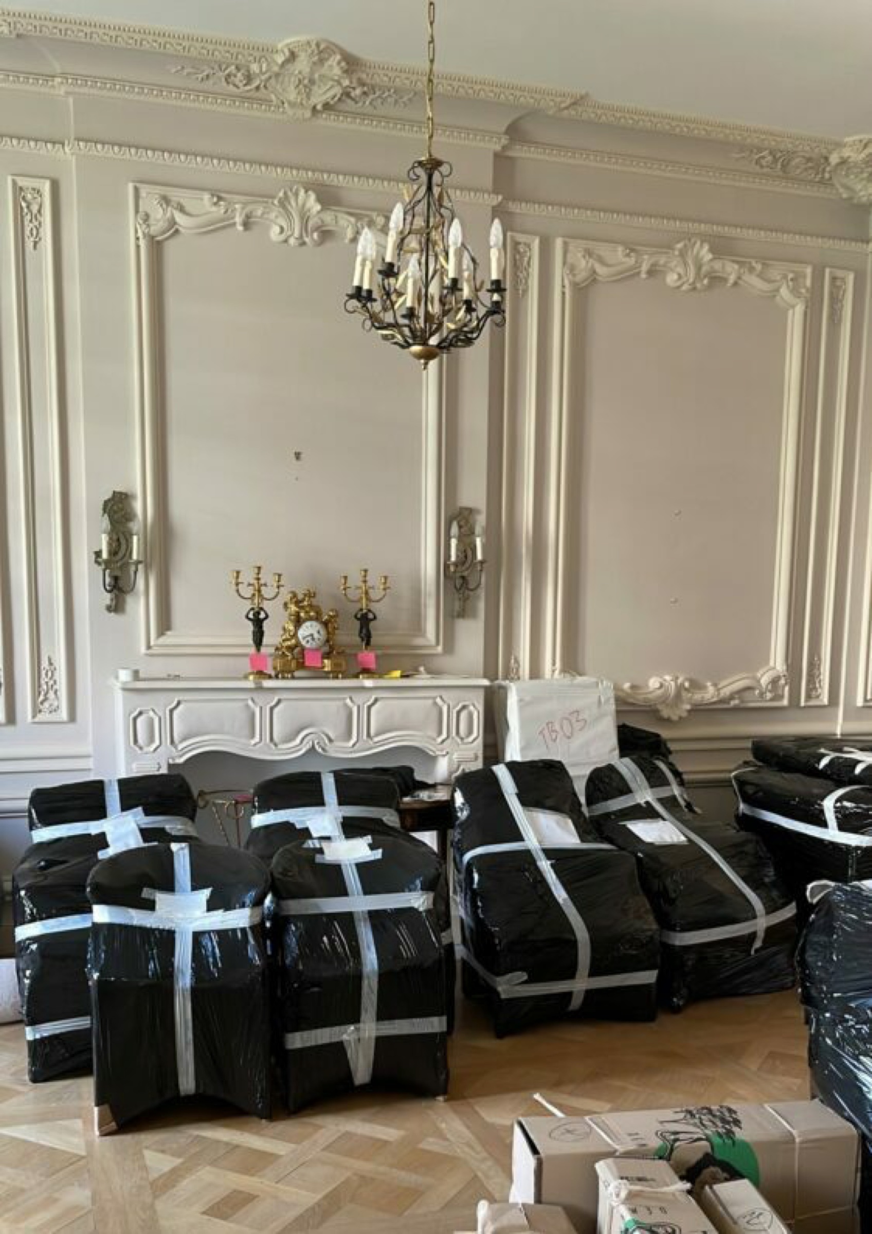 Salon Louis XV avec moulures Régence, lustre cristal et fauteuils emballés sous housse plastique noire Griffon Movers, candélabres bronze sur cheminée